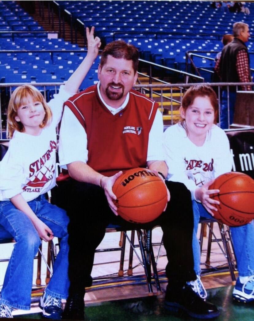 Aly Carr (left) and Cait Carr (far right) sit with their father Chris Carr (center) on the sidelines of a basketball court in 2003. The sisters watched their father coach at various high schools growing up, which later inspired them to become high school basketball coaches themselves. (Photo courtesy of Marla Carr)